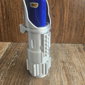 May include: A silver 3D printed lightsaber hilt with a blue BIC lighter inserted. The lighter is visible through the top opening of the hilt.