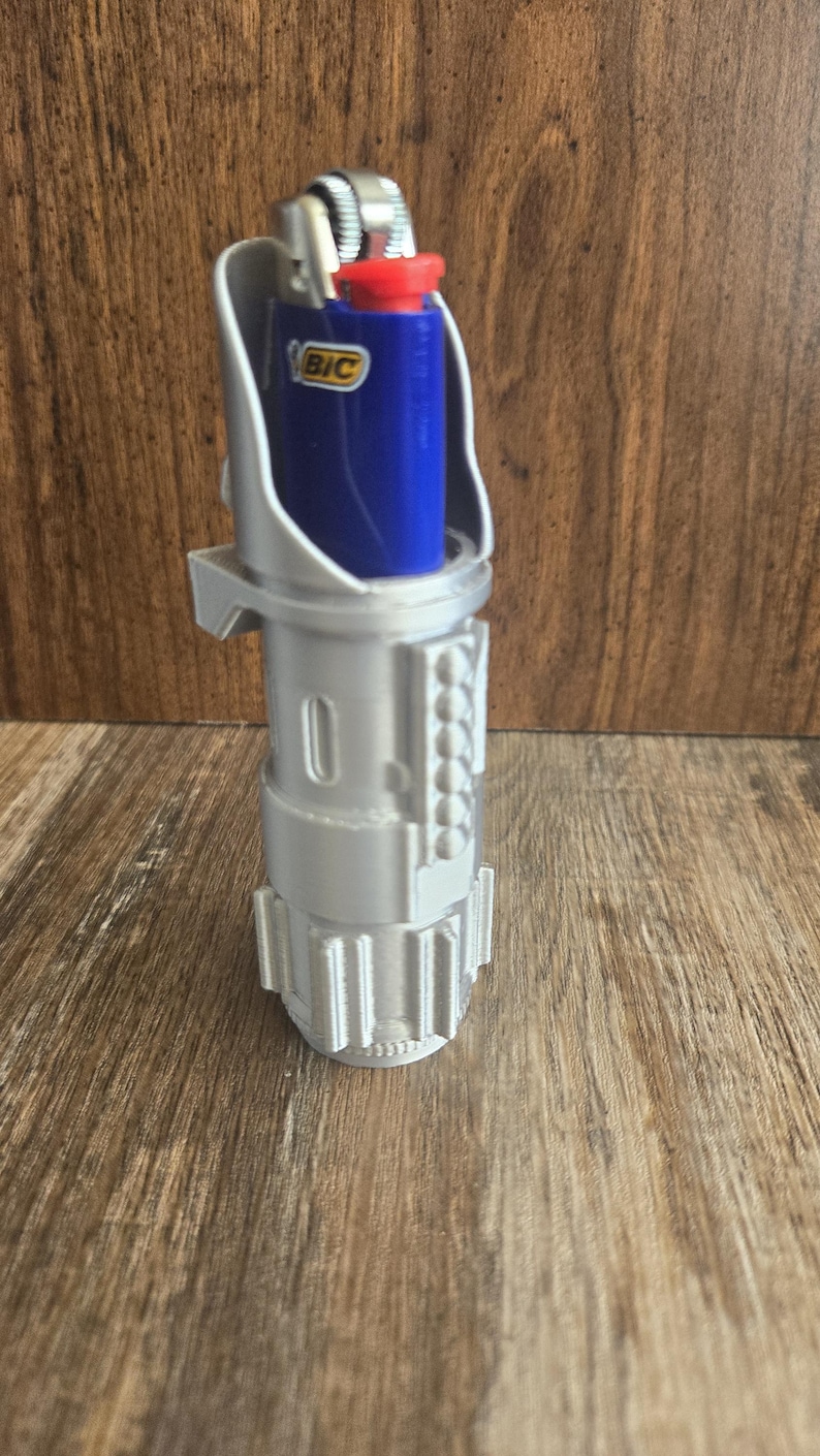 May include: A silver 3D printed lightsaber hilt with a blue BIC lighter inserted. The lighter is visible through the top opening of the hilt.