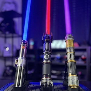 May include: Three illuminated lightsabers with blue, red, and purple blades are displayed on stands. The hilts feature metallic silver, black, and gold finishes. The lightsabers are a popular item in the science fiction genre.