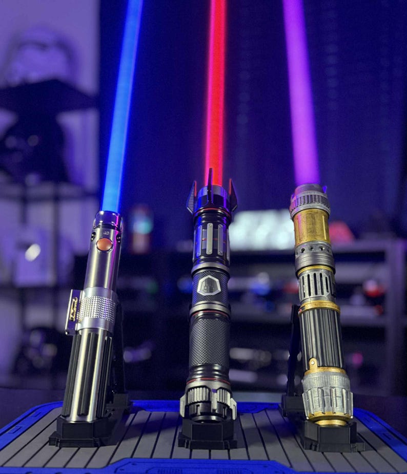 May include: Three illuminated lightsabers with blue, red, and purple blades are displayed on stands. The hilts feature metallic silver, black, and gold finishes. The lightsabers are a popular item in the science fiction genre.