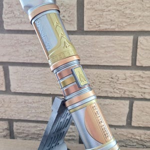 Op de afbeelding: Een zilveren, gouden en koperen lightsaber met ingewikkelde details en de tekst "Seventh Sister" gegraveerd aan de zijkant. De lightsaber staat op een zwarte standaard.