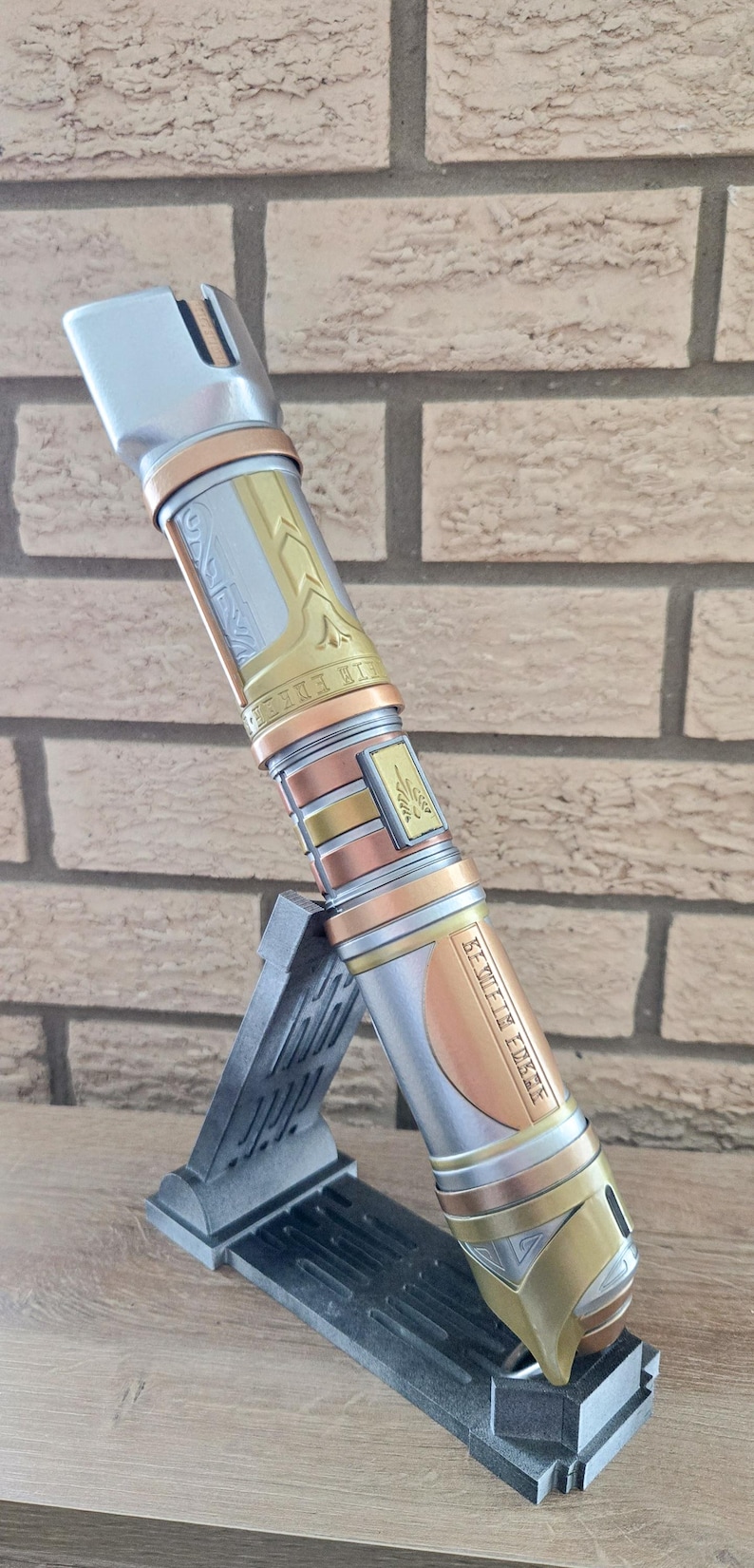 Op de afbeelding: Een zilveren, gouden en koperen lightsaber met ingewikkelde details en de tekst "Seventh Sister" gegraveerd aan de zijkant. De lightsaber staat op een zwarte standaard.
