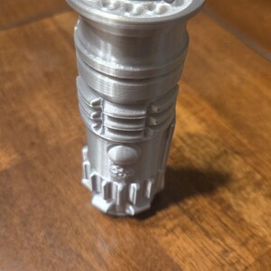 May include: A silver-colored lighter shaped like a lightsaber. The lighter has a red button and a silver top.
