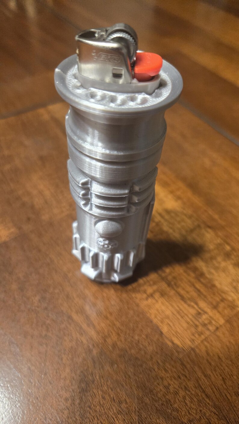 May include: A silver-colored lighter shaped like a lightsaber. The lighter has a red button and a silver top.