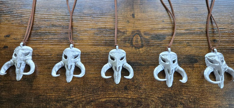 May include: Five identical white skull-shaped pendants with curved horns, each suspended from a thin brown leather cord. The pendants feature hollow eye sockets and a central nasal cavity, creating a distinct, symmetrical, and weathered appearance.