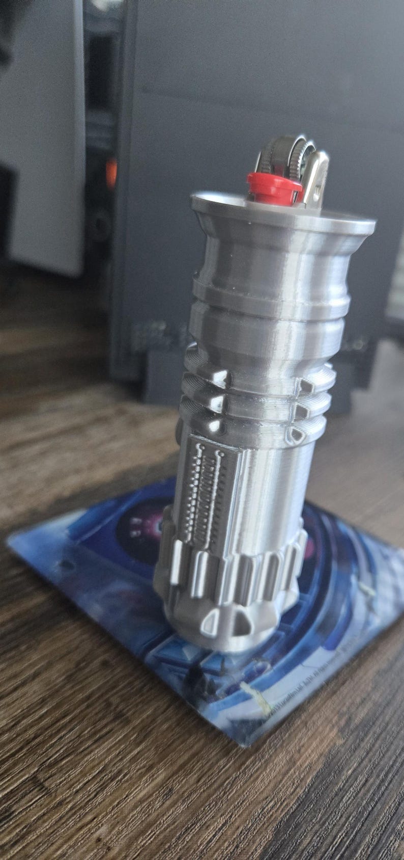 May include: A silver-colored lighter shaped like a lightsaber. The lighter has a red button on the top and a silver metal finish.