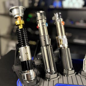 May include: A display of three metallic lightsabers with detailed grips and various button designs. One lightsaber is partially disassembled, revealing internal components. The sabers rest on a blue and gray platform.