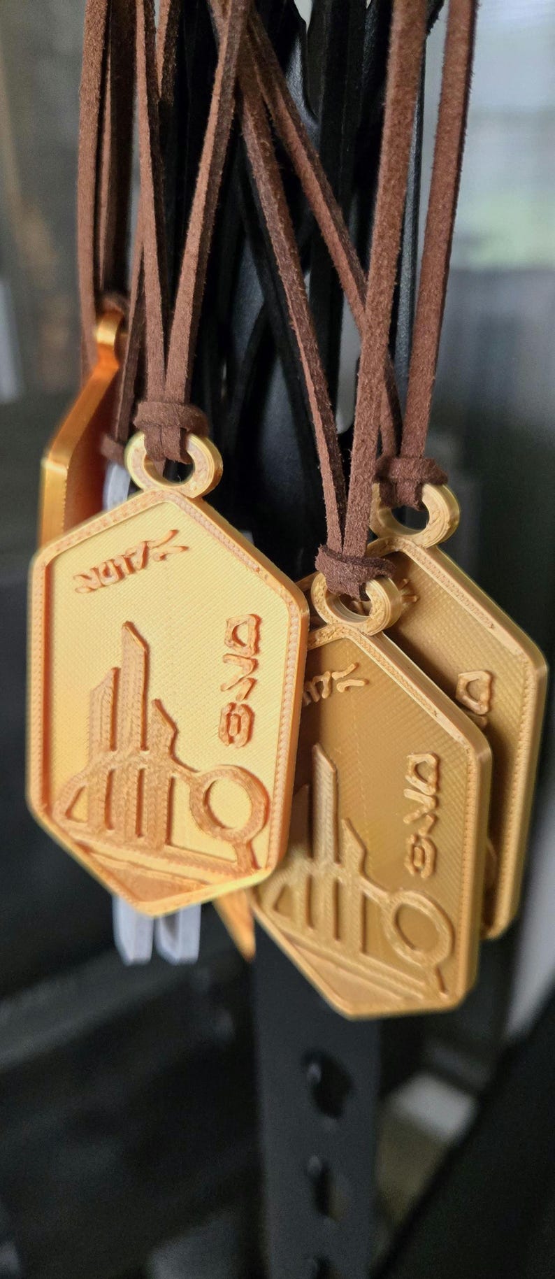 Puede incluir: Collares con colgantes dorados y cordones de cuero marr&oacute;n. Cada colgante tiene un dise&ntilde;o &uacute;nico con elementos arquitect&oacute;nicos y texto. Los colgantes son hexagonales y parecen estar impresos en 3D.