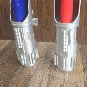 May include: Two silver lightsaber lighters with blue and red lighters inside. The lighters are on a wooden surface.