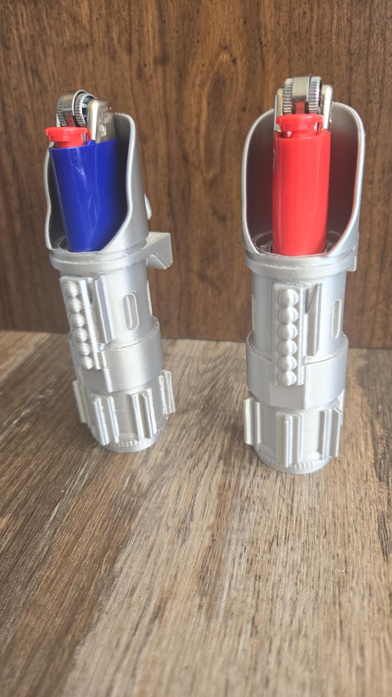 May include: Two silver lightsaber lighters with blue and red lighters inside. The lighters are on a wooden surface.
