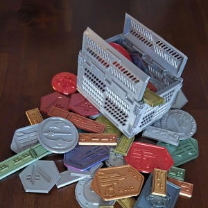 May include: A gray plastic storage box with a lid open, revealing a variety of colorful tokens. The tokens are made of plastic and have different shapes and designs. Some of the tokens have text on them, such as "END" and "SCORE".