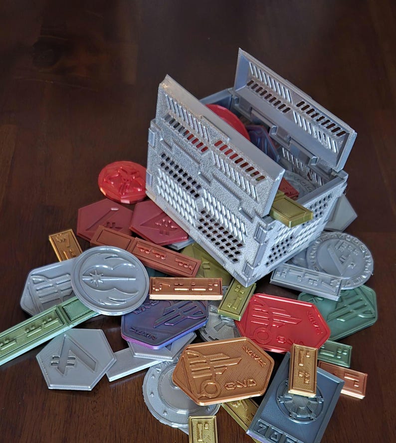 May include: A gray plastic storage box with a lid open, revealing a variety of colorful tokens. The tokens are made of plastic and have different shapes and designs. Some of the tokens have text on them, such as "END" and "SCORE".