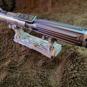 May include: A silver lightsaber with a black handle and a decorative metal plate on the side. The lightsaber is on a white plastic stand.