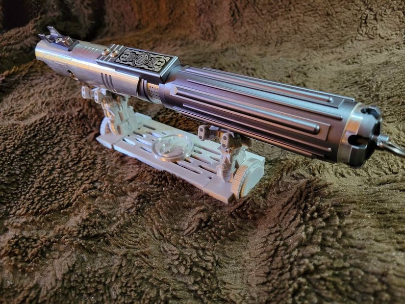 May include: A silver lightsaber with a black handle and a decorative metal plate on the side. The lightsaber is on a white plastic stand.