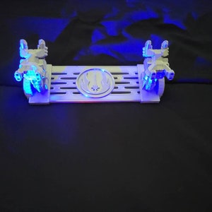 May include: A white plastic display stand with two small, white, detailed model spaceships on either side. The stand has a black and white symbol in the center and a grid pattern on the surface. The stand is lit with blue LED lights.