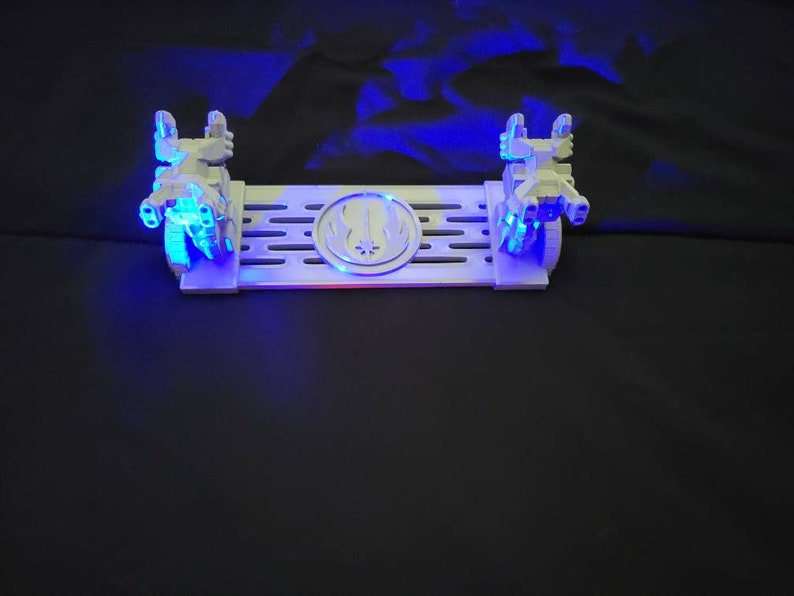 May include: A white plastic display stand with two small, white, detailed model spaceships on either side. The stand has a black and white symbol in the center and a grid pattern on the surface. The stand is lit with blue LED lights.
