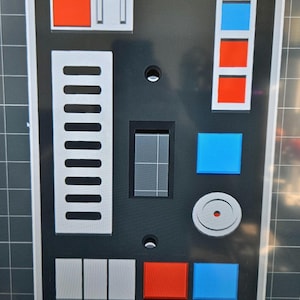 Op de afbeelding: Een zwarte lichtschakelaar-afdekplaat met een Star Wars-thema. Het heeft een reeks knoppen, een schakelaar en een kleine ronde knop met een rode stip in het midden. De knoppen zijn in verschillende kleuren, waaronder rood en blauw.
