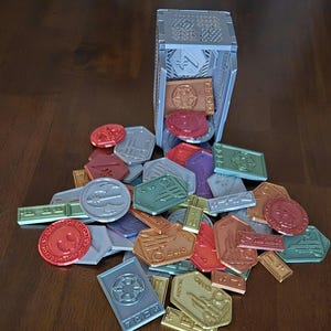 May include: A collection of colorful, metallic tokens with various designs, including a star wars logo, a spaceship, and a lightsaber. The tokens are spilling out of a gray, 3D printed box.