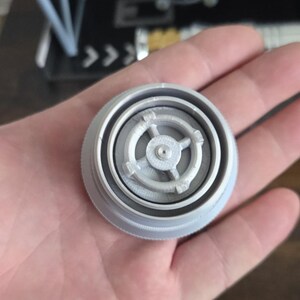 Puede incluir: Un modelo gris claro, impreso en 3D, de un componente de caza TIE. La pieza circular tiene detalles intrincados y se sostiene en una mano. Un modelo de caza TIE m&aacute;s grande es visible en el fondo.
