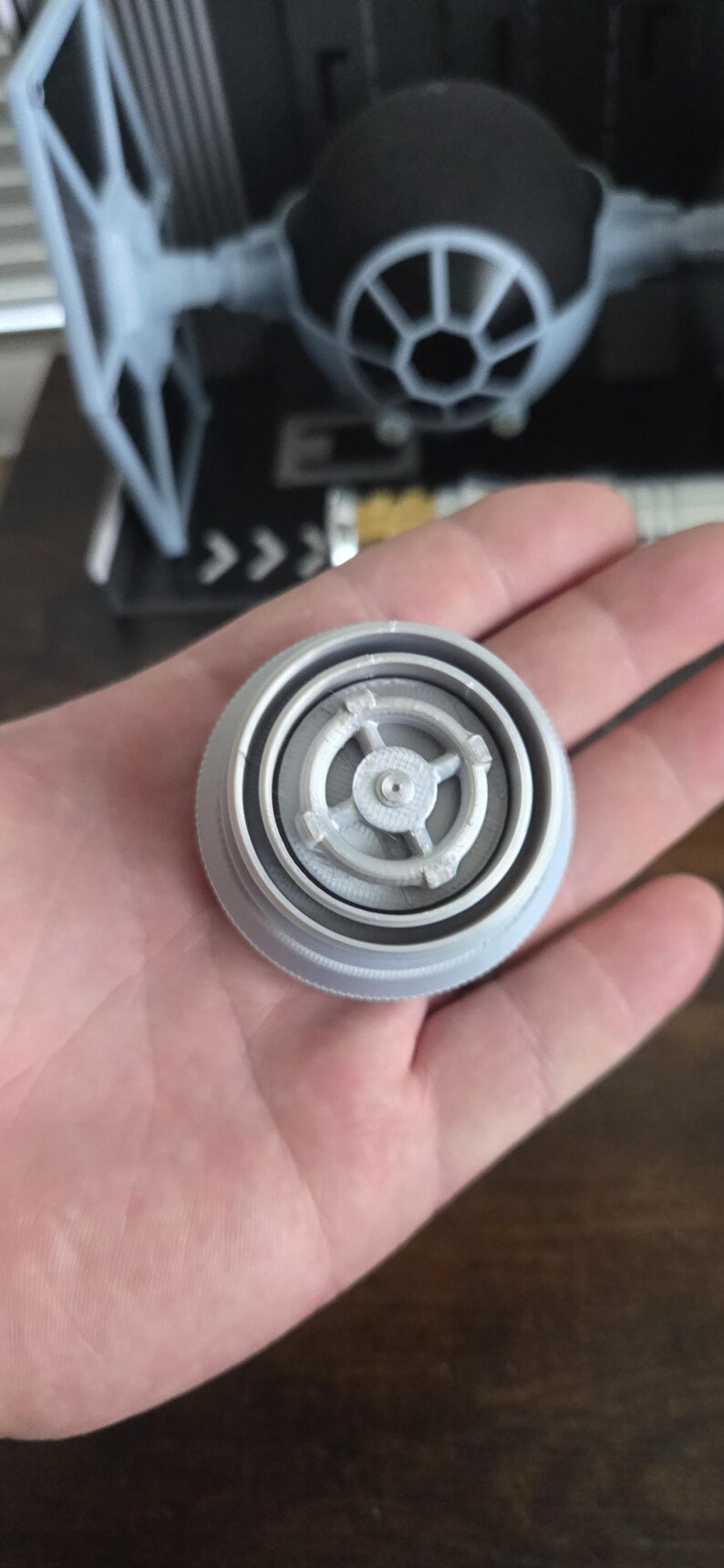 Puede incluir: Un modelo gris claro, impreso en 3D, de un componente de caza TIE. La pieza circular tiene detalles intrincados y se sostiene en una mano. Un modelo de caza TIE m&aacute;s grande es visible en el fondo.