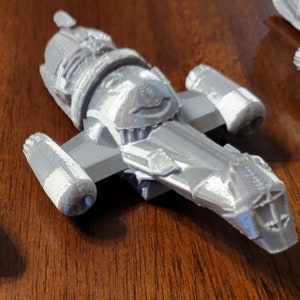May include: A gray 3D printed model of a spaceship with a detailed design. The spaceship has a long, pointed nose and two large engines on the sides.