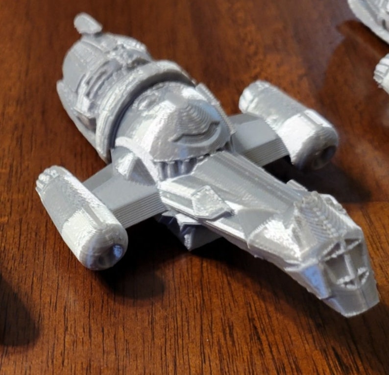 May include: A gray 3D printed model of a spaceship with a detailed design. The spaceship has a long, pointed nose and two large engines on the sides.