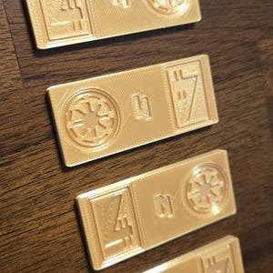Może przedstawiać: Cztery prostokątne, jasnozłote żetony drukowane w 3D. Każdy żeton zawiera symbol, numer i geometryczny wz&oacute;r. Symbole obejmują gwiazdę i stylizowaną literę. Żetony są ułożone na ciemnobrązowej drewnianej powierzchni.