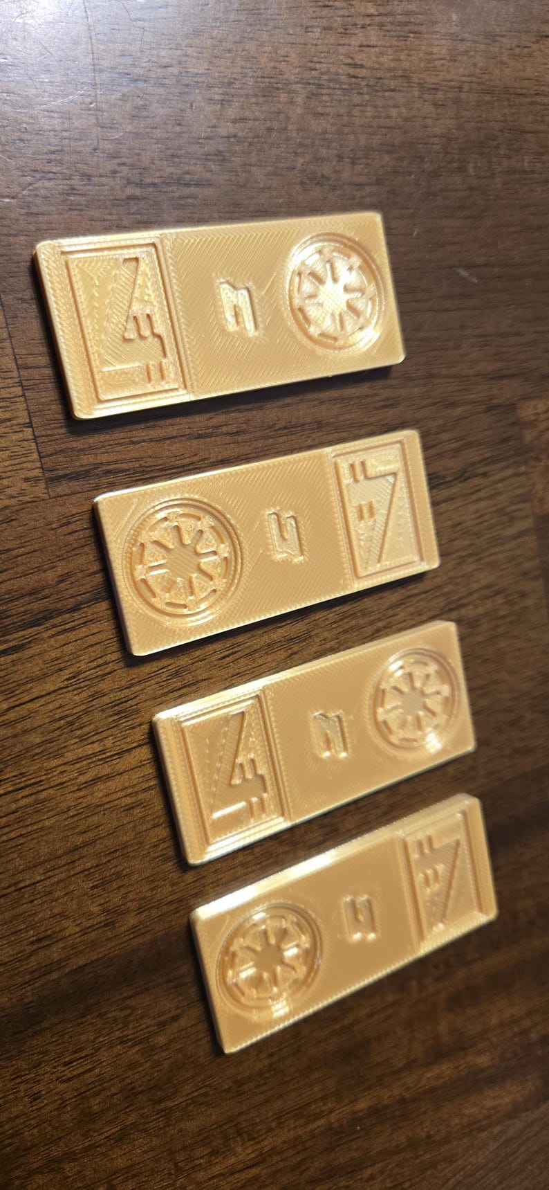 Może przedstawiać: Cztery prostokątne, jasnozłote żetony drukowane w 3D. Każdy żeton zawiera symbol, numer i geometryczny wz&oacute;r. Symbole obejmują gwiazdę i stylizowaną literę. Żetony są ułożone na ciemnobrązowej drewnianej powierzchni.