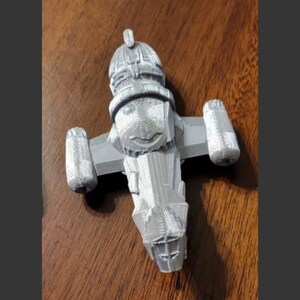 May include: A 3D printed model of a gray spaceship with a rounded top and two wings. The spaceship has a detailed design with a textured surface.