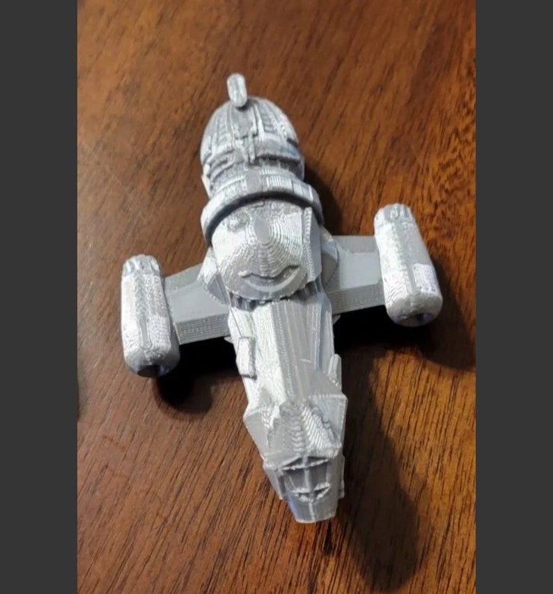 May include: A 3D printed model of a gray spaceship with a rounded top and two wings. The spaceship has a detailed design with a textured surface.