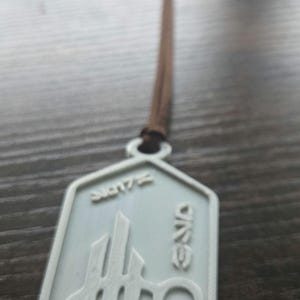 Puede incluir: Un colgante gris claro con un cord&oacute;n marr&oacute;n. El colgante tiene un dise&ntilde;o geom&eacute;trico con la palabra "ONE" y el n&uacute;mero "2017". El colgante est&aacute; hecho de un material gris claro.