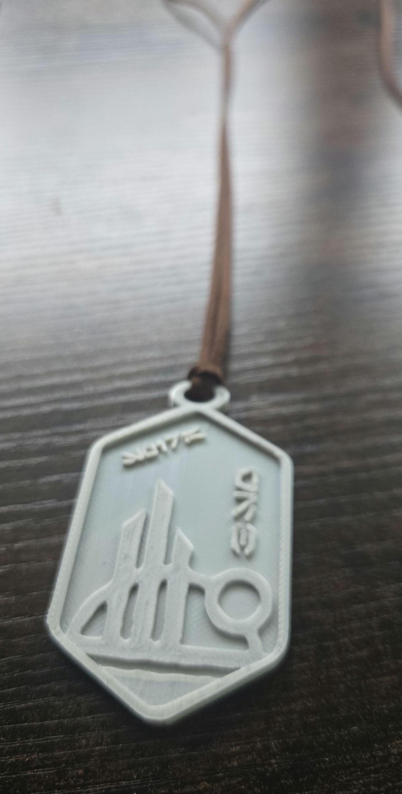 Puede incluir: Un colgante gris claro con un cord&oacute;n marr&oacute;n. El colgante tiene un dise&ntilde;o geom&eacute;trico con la palabra "ONE" y el n&uacute;mero "2017". El colgante est&aacute; hecho de un material gris claro.