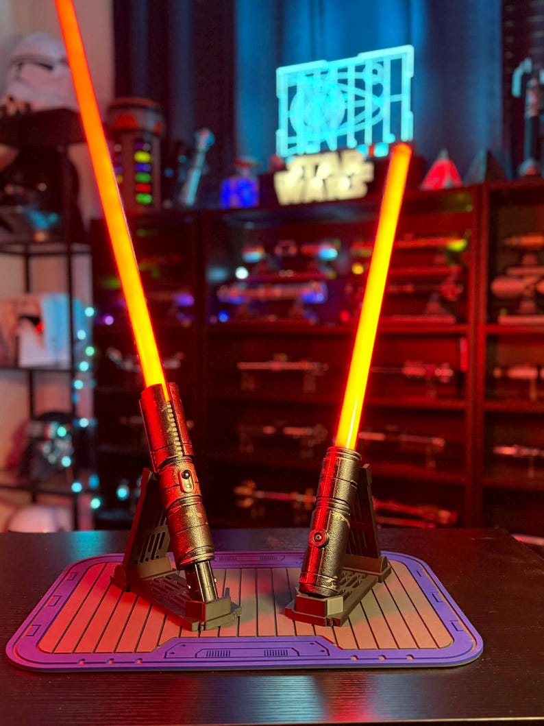 K&ouml;nnte beinhalten: Zwei Lichtschwerter mit leuchtend orangefarbenen Klingen. Die metallischen Griffe sind detailliert und stehen auf einzelnen St&auml;ndern. Die Pr&auml;sentation befindet sich auf einer lila-roten Matte. Der Hintergrund zeigt ein "Star Wars"-Schild und andere Sammlerst&uuml;cke.