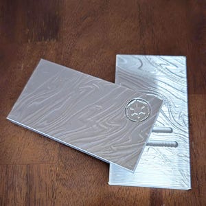 May include: Two silver-coloured, rectangular, 3D printed plates with a textured surface. One plate has a raised, circular design with a star in the centre. The other plate has three rectangular slots.