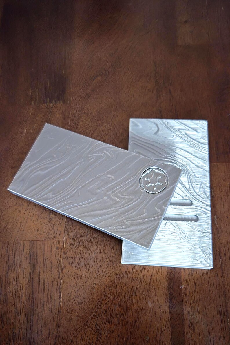May include: Two silver-coloured, rectangular, 3D printed plates with a textured surface. One plate has a raised, circular design with a star in the centre. The other plate has three rectangular slots.