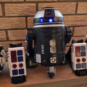 Peut inclure: Une collection de jouets de dro&iuml;des Star Wars, dont BB-8, R2-D2 et d'autres robots sph&eacute;riques et cylindriques. Les dro&iuml;des sont noirs, blancs et argent&eacute;s, avec divers d&eacute;tails et antennes. Ils sont expos&eacute;s sur une surface en bois.
