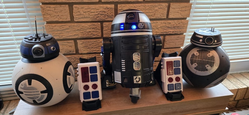 Peut inclure: Une collection de jouets de dro&iuml;des Star Wars, dont BB-8, R2-D2 et d'autres robots sph&eacute;riques et cylindriques. Les dro&iuml;des sont noirs, blancs et argent&eacute;s, avec divers d&eacute;tails et antennes. Ils sont expos&eacute;s sur une surface en bois.