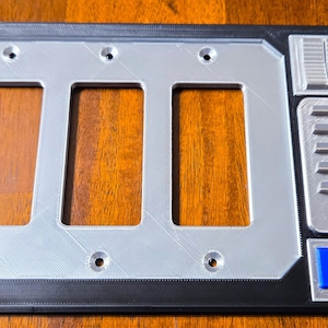 May include: A rectangular light switch plate with three rectangular openings, a silver-colored frame, and a black border. The plate has a design with various shapes and colors, including blue and orange squares and an orange circle.