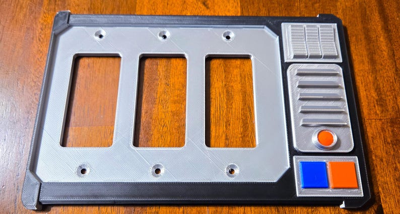May include: A rectangular light switch plate with three rectangular openings, a silver-colored frame, and a black border. The plate has a design with various shapes and colors, including blue and orange squares and an orange circle.