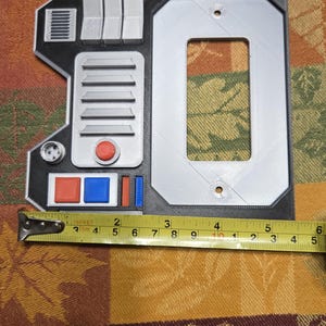 May include: A 3D printed wall plate with a black and gray design. The plate has a rectangular opening for a light switch and features a small, round, red button, a red and blue button, and a small, round, gray knob. The plate is 4 inches wide.