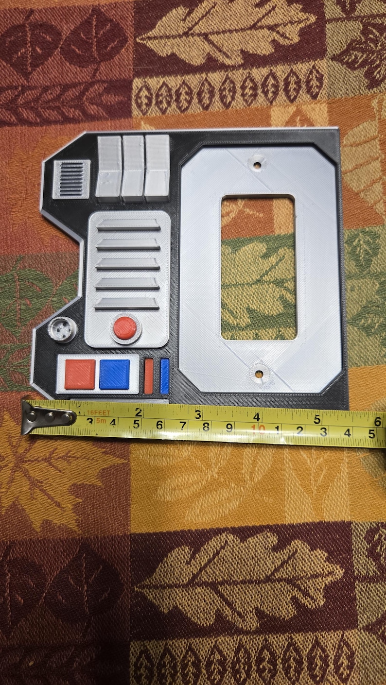 May include: A 3D printed wall plate with a black and gray design. The plate has a rectangular opening for a light switch and features a small, round, red button, a red and blue button, and a small, round, gray knob. The plate is 4 inches wide.