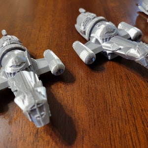 May include: Three gray 3D printed model spaceships, each with a distinct design, are displayed on a wooden surface.