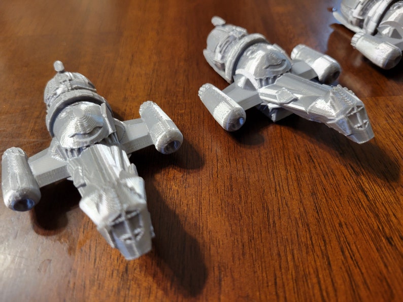May include: Three gray 3D printed model spaceships, each with a distinct design, are displayed on a wooden surface.
