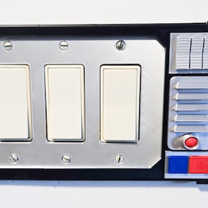 May include: A unique light switch cover with a retro-futuristic design. It features three white light switches, a silver-colored faceplate, and a black border. Additional details include a red button, blue and red squares, and a series of gray vents.