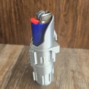May include: A silver 3D printed lighter holder shaped like a lightsaber. A blue lighter is inserted into the holder.