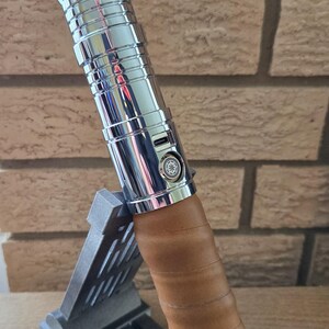 Op de afbeelding: Een zilveren en gouden lightsaber met een bruine leren grip, rustend op een grijze standaard. De lightsaber heeft een zilveren body met gouden accenten en een kleine zilveren knop aan de zijkant. De leren grip is om de lightsaber gewikkeld en heeft een bruine kleur.