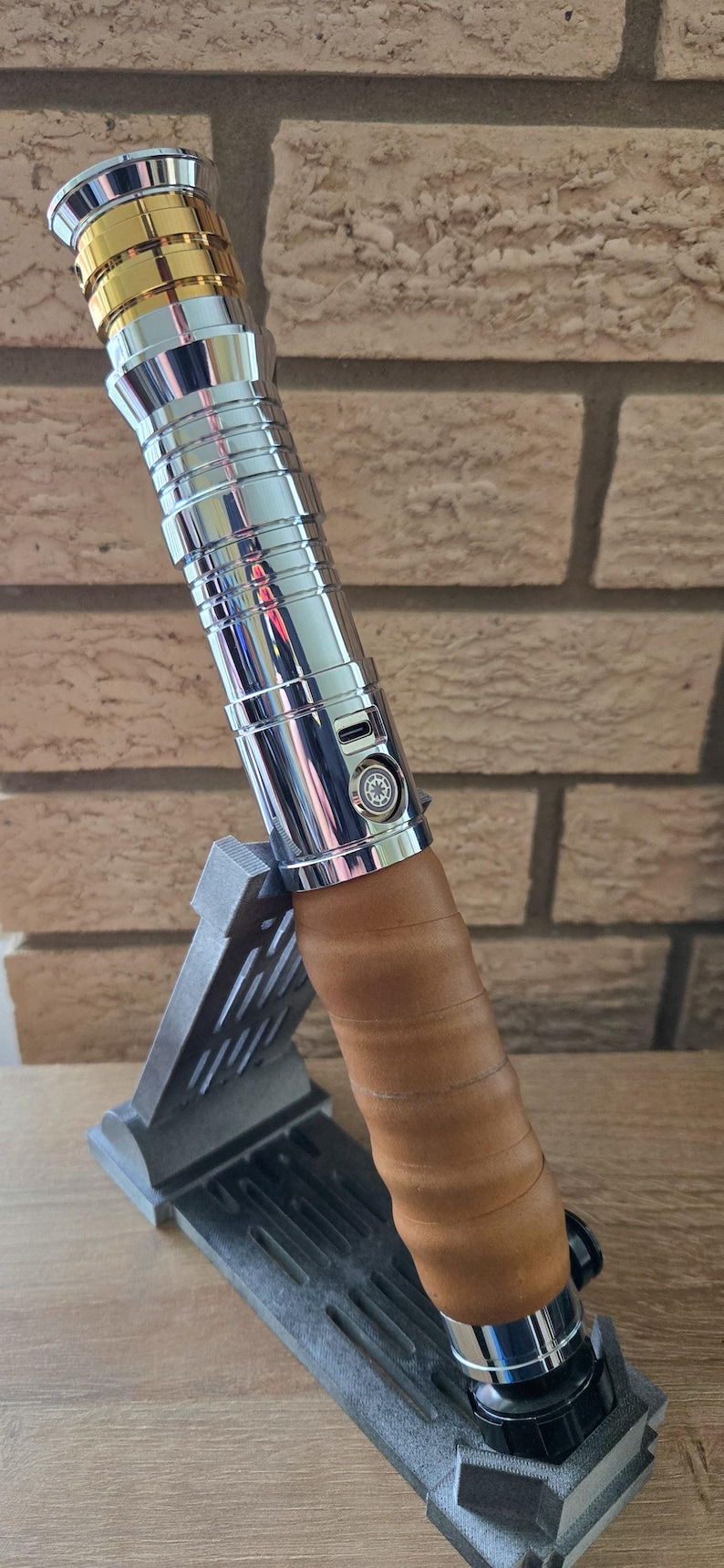 Op de afbeelding: Een zilveren en gouden lightsaber met een bruine leren grip, rustend op een grijze standaard. De lightsaber heeft een zilveren body met gouden accenten en een kleine zilveren knop aan de zijkant. De leren grip is om de lightsaber gewikkeld en heeft een bruine kleur.