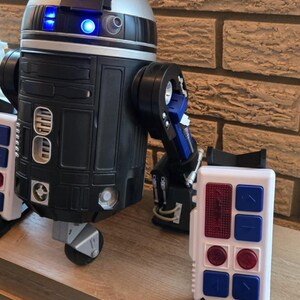 Peut inclure: Un jouet robot noir, argent&eacute; et bleu avec un panneau de commande blanc. Le robot a une t&ecirc;te cylindrique avec des lumi&egrave;res bleues et un corps rectangulaire. Le panneau de commande a des boutons rouges et bleus.
