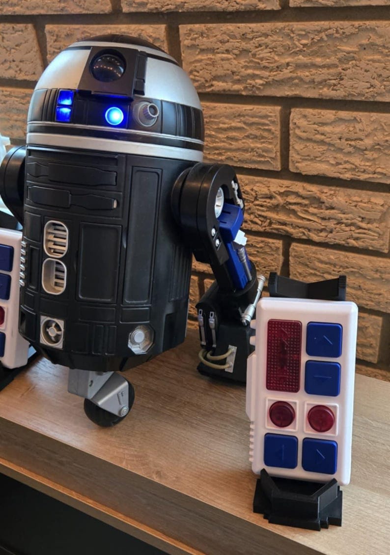 Peut inclure: Un jouet robot noir, argent&eacute; et bleu avec un panneau de commande blanc. Le robot a une t&ecirc;te cylindrique avec des lumi&egrave;res bleues et un corps rectangulaire. Le panneau de commande a des boutons rouges et bleus.