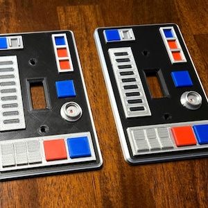 Op de afbeelding: Twee zwarte lichtschakelaars met een zilveren en grijs ontwerp. De afdekkingen hebben verschillende knoppen en schakelaars, waaronder rode en blauwe knoppen. De afdekkingen zijn ontworpen om eruit te zien als een bedieningspaneel van een ruimteschip.